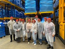 Gruppe von Personen mit roten Haarnetzen und weißen Schutzkitteln steht in einem Lager mit hohen Regalen und blauen Paletten