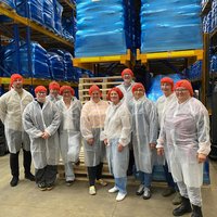 Gruppe von Personen mit roten Haarnetzen und weißen Schutzkitteln steht in einem Lager mit hohen Regalen und blauen Paletten
