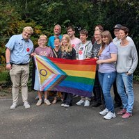 Gründungsmitglieder vom Queeren Netzwerk Bramsche stehen im Freien vor Büschen und halten die aktualisierte Progress-Flag