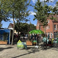 Das Bild zeigt den Bramscher Marktplatz. Im Mittelpunkt steht ein Informationsstand der Partei Bündnis 90/Die Grünen. Ein umfunktionierter Container, die Klimakiste dient als Hintergrund, Tische und Stühle laden zum Verweilen ein, Fahrräder stehen in der Nähe. Ein Banner mit der Aufschrift "Es gibt keinen Planet B" unterstreicht den ökologischen Fokus der Veranstaltung. Die Szene wirkt ruhig und sonnig, die Atmosphäre entspannt. Im Hintergrund sind weitere Gebäude und Bäume zu sehen.