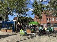Das Bild zeigt den Bramscher Marktplatz. Im Mittelpunkt steht ein Informationsstand der Partei Bündnis 90/Die Grünen. Ein umfunktionierter Container, die Klimakiste dient als Hintergrund, Tische und Stühle laden zum Verweilen ein, Fahrräder stehen in der Nähe. Ein Banner mit der Aufschrift "Es gibt keinen Planet B" unterstreicht den ökologischen Fokus der Veranstaltung. Die Szene wirkt ruhig und sonnig, die Atmosphäre entspannt. Im Hintergrund sind weitere Gebäude und Bäume zu sehen.