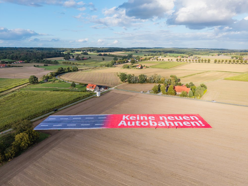 Luftaufnahme eines riesigen Banners auf einem Feld. Die eine häfte sind vier Fahrstreifen und die andere hat die Aufschrift 'Keine neuen Autobahnen!' in weißer Schrift auf rotem Hintergrund