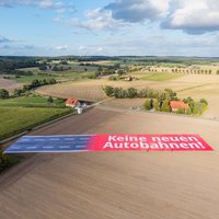 Luftaufnahme eines riesigen Banners auf einem Feld. Die eine häfte sind vier Fahrstreifen und die andere hat die Aufschrift 'Keine neuen Autobahnen!' in weißer Schrift auf rotem Hintergrund