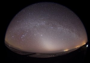 Weitwinkelaufnahme eines Sternenhimmels mit sichtbarer Lichtemission am Horizont über einer dunklen Landschaft