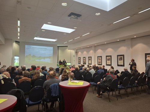 Vortragsraum mit Publikum bei der Tagung Naturschutz des Umweltforums Osnabrück 2026, Personen sitzen vor einer Leinwand mit Präsentation, an der Wand hängen gerahmte Bilder