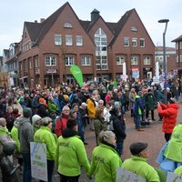 Menschenkette auf dem Münsterplatz in Bramsche mit vielen Teilnehmenden, einige tragen grüne Jacken mit der Aufschrift 'amal' und halten Schilder, im Hintergrund Backsteingebäude und eine Fahne von Bündnis 90/Die Grünen