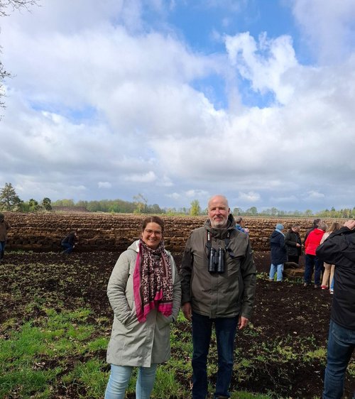 Zwei Personen stehen auf einem Moorboden, im Hintergrund weitere Menschen und ein bewölkter Himmel