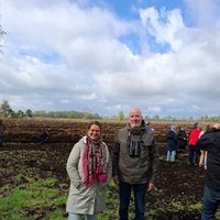 Zwei Personen stehen auf einem Moorboden, im Hintergrund weitere Menschen und ein bewölkter Himmel