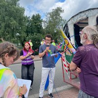Fabian Sommer (Kreisverbandssprecher Grüne) spricht mit Mikrofon vor einer kleinen Gruppe beim CSD Bramsche, umgeben von Personen mit Warnwesten und Fahnen im Hintergrund