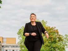 Frau (Mareen Guth) in schwarzer Kleidung geht auf einer Straße mit Gebäuden und Bäumen im Hintergrund