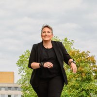 Frau (Mareen Guth) in schwarzer Kleidung geht auf einer Straße mit Gebäuden und Bäumen im Hintergrund