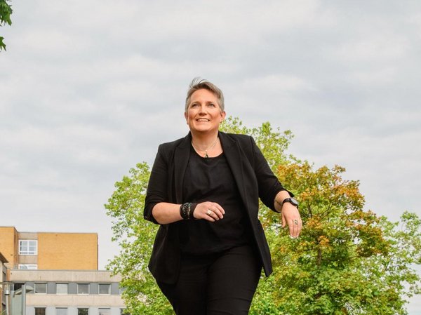 Frau (Mareen Guth) in schwarzer Kleidung geht auf einer Straße mit Gebäuden und Bäumen im Hintergrund