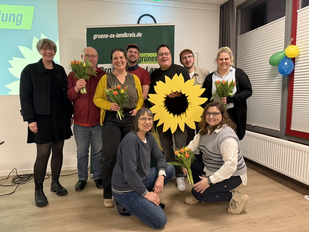 Gruppenbild des neuen und alten Vorstandes, einige stehend, einige kniend, halten Blumen und ein großes gelbes Sonnenblumen-Symbol 