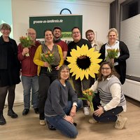 Gruppenbild des neuen und alten Vorstandes, einige stehend, einige kniend, halten Blumen und ein großes gelbes Sonnenblumen-Symbol 